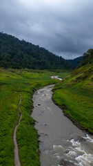 Clear mountain water flows over rocks in a vibrant green valley, surrounded by majestic peaks at dawn.