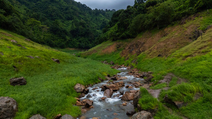 Clear mountain water flows over rocks in a vibrant green valley, surrounded by majestic peaks at dawn.