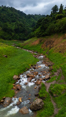 Clear mountain water flows over rocks in a vibrant green valley, surrounded by majestic peaks at dawn.