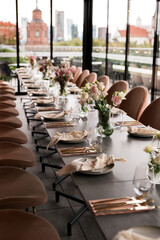 Elegant long table setting with flowers, gold cutlery, and neutral-toned decor in a modern restaurant with panoramic city views, prepared for a wedding, event, or upscale dining experience.