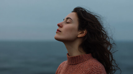 A young woman stands by the sea, embracing tranquility with closed eyes and wind-swept hair.