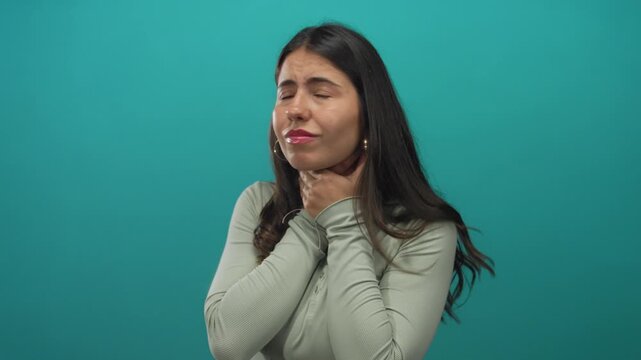 Young hispanic woman holds throat with both hands for a sore throat in a green studio; health concern discomfort.