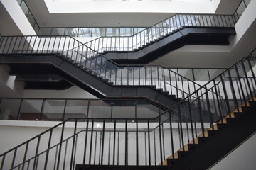 Modern multi-level atrium with sleek black metal staircases, glass railings, and wooden steps. Bright, open space with red seating and minimalist design.