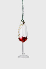 Minimal creative aesthetic view of Glass of red wine Christmas decoration hanging on grey. New Year bright glass decor as wineglass. Festive minimal concept of celebration, party and winter holidays.