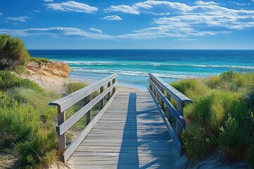 Wooden boardwalk through green sand dunes leading to a turquoise ocean with gentle waves and a rocky outcrop under a bright blue sky with scattered clouds, serene and inviting