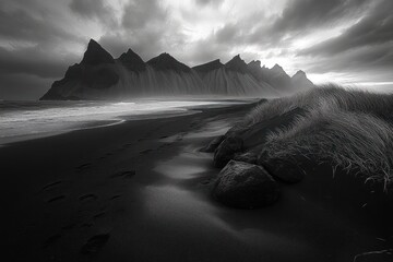 moody black sand beach with jagged mountain silhouettes, footprints along the shoreline past grassy dunes and rocks beneath a dramatic stormy sky