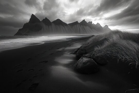 moody black sand beach with jagged mountain silhouettes, footprints along the shoreline past grassy dunes and rocks beneath a dramatic stormy sky