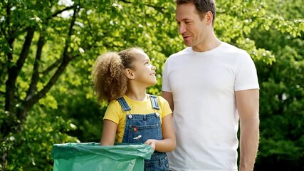 A father and daughter smile while holding a green trash bag outdoors. - Powered by Adobe