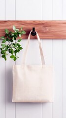 A plain, light-colored canvas tote bag hangs on a dark hook attached to a wooden plank, with green leaves on the left.