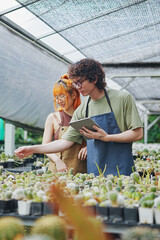 Young Asian Man and Woman Gardeners Working Together in Greenhouse with Tablet, Caring for Cactus Flowers and Plants
