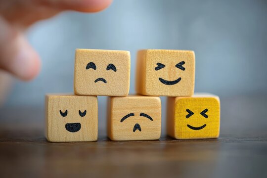 Hand reaching toward five small wooden blocks painted with simple faces showing mixed emotions — smiling, laughing, sad and worried, conveying a playful and reflective mood