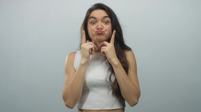 Young woman with long dark hair wearing a white tank top pointing fingers to her cheeks in a gray studio; playful.