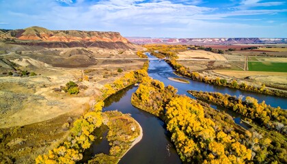 Aerial view of river winding through arid landscape with golden trees and distant mesas under a bright blue sky