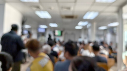 Blurred image depicting the interior of busy hospital waiting room or clinic lobby. Numerous people are seated and standing, suggesting high volume of patients