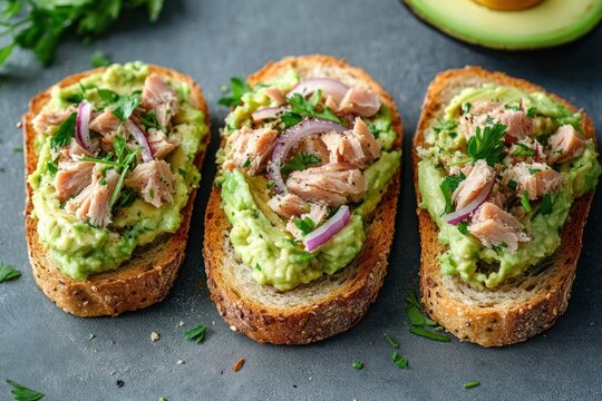 Three slices of rustic multigrain bread topped with mashed avocado, flaked fish, red onion slices and parsley, fresh and appetizing