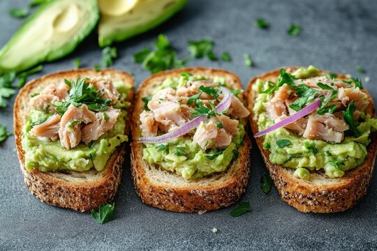 Three slices of seeded toast topped with mashed avocado, flaked tuna, red onion and parsley on a dark surface with avocado halves in the background, fresh and appetizing brunch