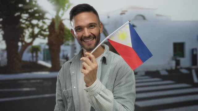 Young man smiling while holding a philippine flag on an urban city street, showcasing cultural pride in a vibrant cityscape background outdoors.