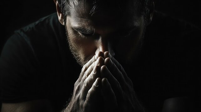 Low-key portrait of a man in a black shirt with clasped hands and tense forearms, dramatic side lighting and deep shadows conveying solemn contemplation and quiet hope