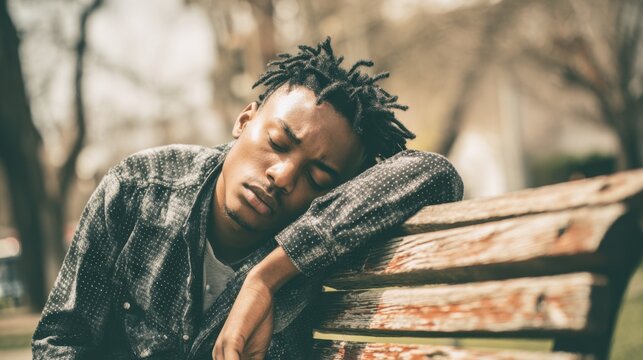 A young man with dreadlocks is sitting on a wooden bench in a park, resting his head on his arm while looking peaceful. It is a sunny day with trees in the background. - Powered by Adobe