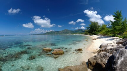 Serene tropical beach features clear turquoise waters and a rocky shoreline. Lush green mountains rise in the background under a bright blue sky filled with fluffy clouds.