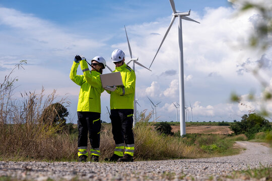 Wind farm technicians inspect equipment and analyze data in bright safety gear under a clear sky near wind turbines