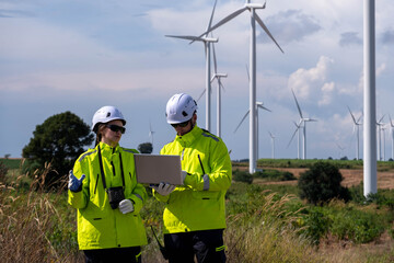 Workers collaborate on a laptop while monitoring wind turbines in a renewable energy project in a sunny landscape