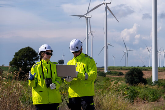 Workers collaborate on a laptop while monitoring wind turbines in a renewable energy project in a sunny landscape