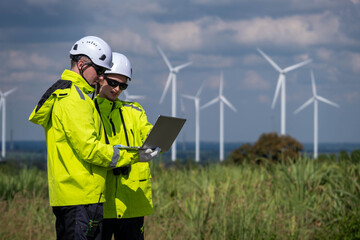 Two workers in bright gear check data on a laptop near wind turbines in a green field on a clear day