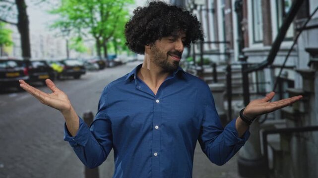 Man with curly hair smiling, palms up in an open hands shrug on a tree lined city street with parked cars and row houses; playful.