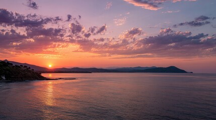 Fototapeta premium Vibrant sunset paints the sky with hues of orange and purple as the sun sets over a peaceful sea. Distant mountains create a stunning backdrop, enhancing the tranquil evening ambiance.