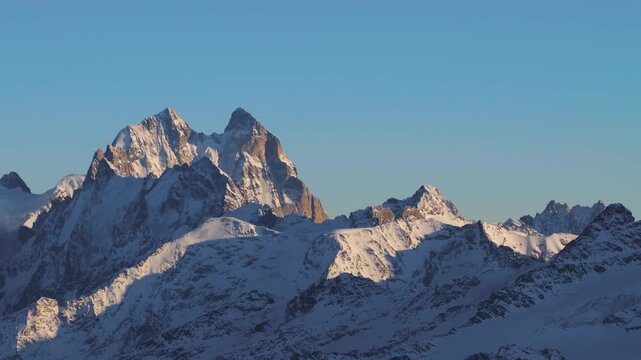 A view of the snow capped Ushba mountain peak, part of the Greater Caucasus range, under a clear blue sky. The footage captures the stunning natural beauty and rugged landscape of the North Caucasus r