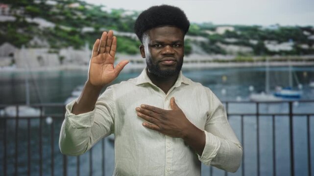 Man with hand on chest and raised palm to camera on street marina with boats and waterfront railing; sincere pledge.