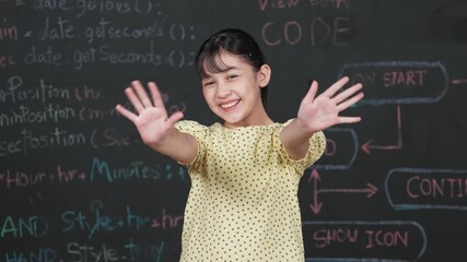 Cute happy girl waving to camera while standing at blackboard with engineering prompt or coding, programming system written in STEM technology classroom. Smiling student looking at camera. Edification