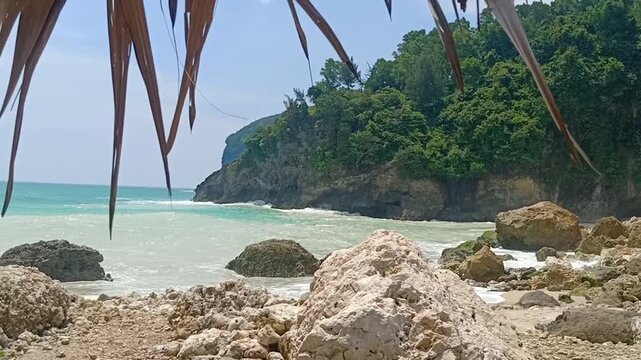 Calm waves sweep across the rocky shore of Sawangan Beach, Kebumen, creating a peaceful coastal view. Turquoise water, scattered limestone rocks, and green cliff combine to form a soothing and refresh
