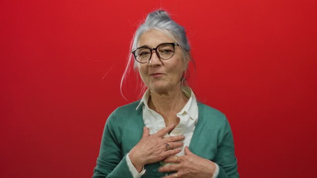 Senior woman with grey hair and glasses in green sweater poses against a vibrant red background, expressing gratitude with hands on chest, radiating warmth and confidence.
