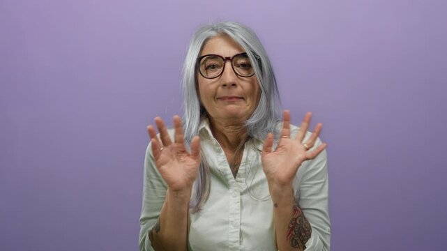Woman with grey hair expressing surprise isolated against a purple background, wearing glasses and white shirt, conveying senior emotions in an isolated studio environment.