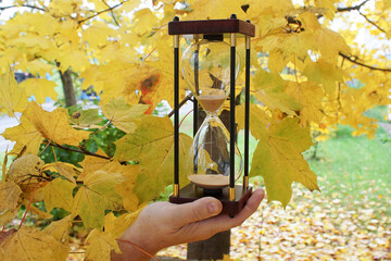 An hourglass in a hand against the backdrop of a tree with yellow leaves. The hourglass as a symbol of time. The concept of time passing irrevocably.