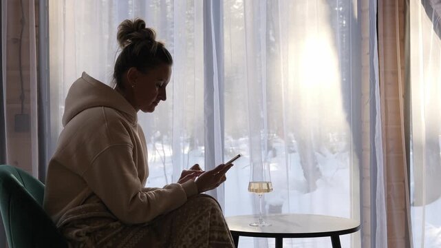 Woman enjoying mindful evening in cozy cabin holding wine and phone near window Phone symbolizes connection reflection gentle balance of offline peace and online world Calm phone moment of awareness