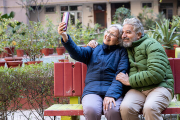 Happy indian senior couple sitting together outdoor in winter season taking selfie picture with smart phone wearing wearing warm clothes. Retirement lifestyle. Active elderly husband wife bonding.