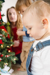 A toddler stands in focus by a Christmas tree, with family decorating in the background.
