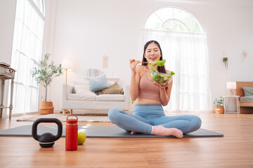 Fototapeta premium Healthy life concept with Asian woman in fitness on yoga mat and eating vegetable salad at home. clean living, nutritional balance, and mindful routine.