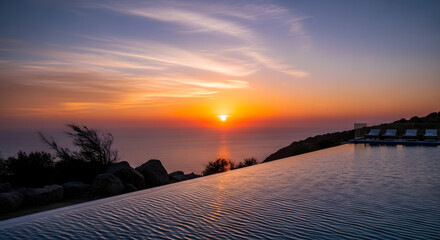 Infinity pool reflecting a vibrant sunset over the ocean with a rocky coastline view and lounge chairs