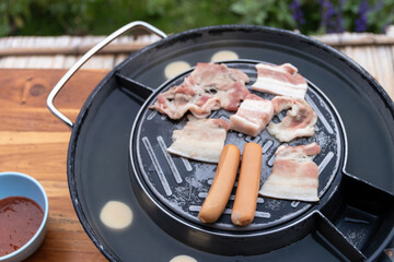 Grilled bacon slices and two sausages on tabletop barbecue pan with dipping sauce, casual outdoor meal and savory aroma