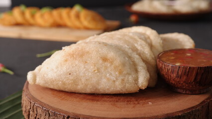 White crescent-shaped pastries served with a red dipping sauce on a rustic wooden board, with more food in the blurred background.