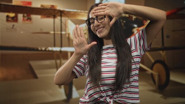 Woman frames face with hands in front of a vintage airplane exhibit inside a building, wearing glasses and striped top; joyful curiosity.