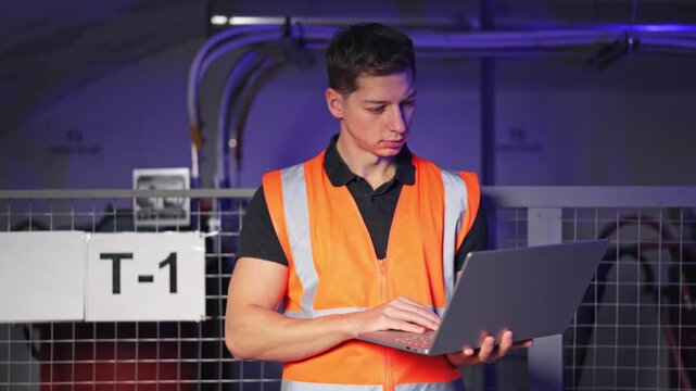 Worker electrician in safety vest opens and types on laptop, focused on operations within an industrial environment. Diligent task completion for maintenance or inventory control.