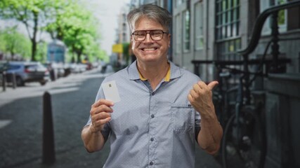 Man holding a creditcard in his right hand, thumb pointing right while smiling on a street with parked cars and a bicycle rack; confidence.