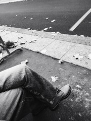 Crossed Legs on a Sidewalk. A person sits with legs crossed on a city sidewalk at the bus stop, surrounded by fallen leaves. The monochrome color scheme adds a calm, reflective atmosphere.
