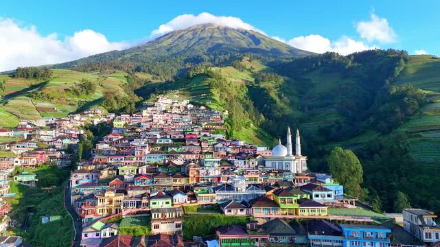 Aerial view of Nepal van Java village nestled on the lush slopes of Mount Sumbing, Indonesia. The colorful houses and terraced hills create a stunning mountain landscape under a bright blue sky.