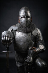 A portrait of a medieval knight in full plate armor and helmet, holding a sword and kneeling, isolated against a dark background. The warrior is kneeling.